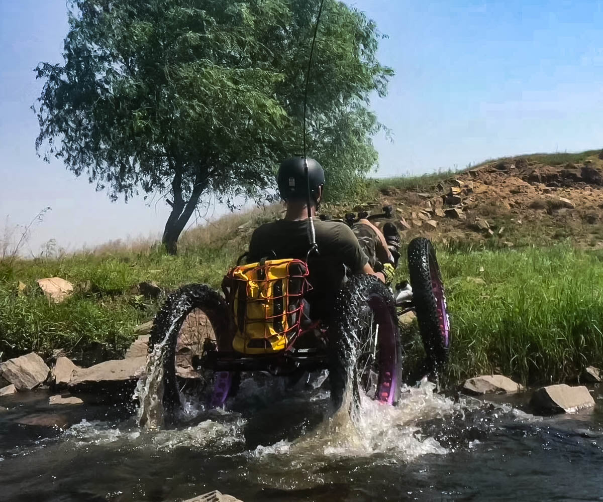 A rider on an FT26e off-road recumbent trike crosses a rocky stream, with water splashing and a tree on a grassy hill in the background.