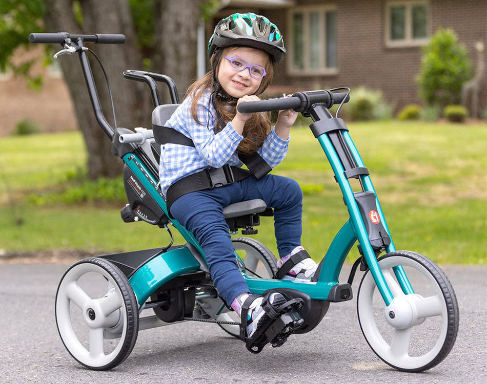 Child riding a Rifton X320 with a teal frame and backrest, wearing a helmet and smiling, in an outdoor setting.