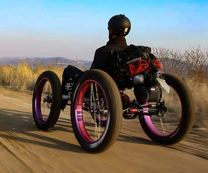 A rider on a FT26e off-road recumbent quad travels down a dirt path with a scenic backdrop of fields and distant hills at sunset