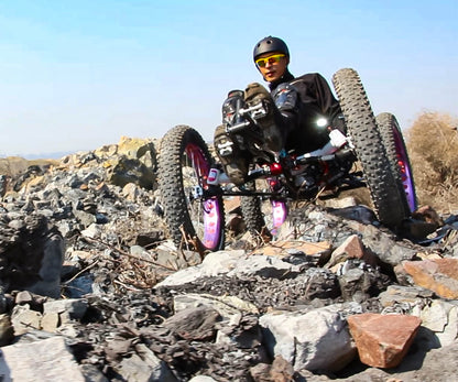 A rider in a helmet and glasses seated at an angle while navigating very rocky terrain on an FT26e off-road recumbent trike